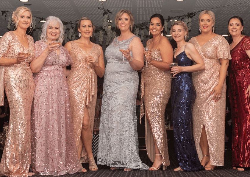 A row of eight women in glitzy ball gowns holding champagne