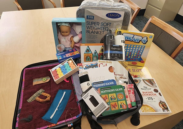 A kit containing a weighted blanket, bluetooth speaker, shampoo, hair brush, hand cream, toy doll, connect 4 game and puzzle books