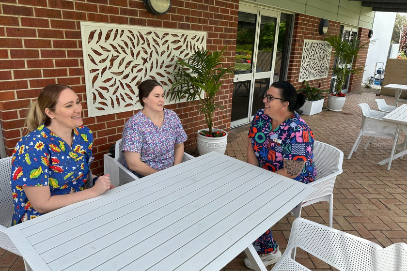 3 ladies in colourful scrubs are talking over a table while sitting outside of a brick building