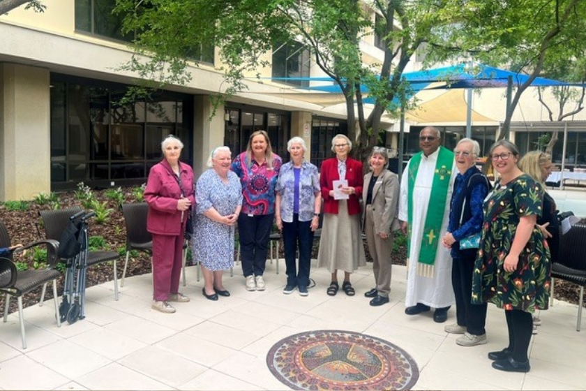 A group of older adults and a priest stand together in a leafy outdoor courtyard beside a building, smiling for a photo. A decorative mosaic is set into the tiled ground in front of them, and chairs and a walking frame are visible nearby.