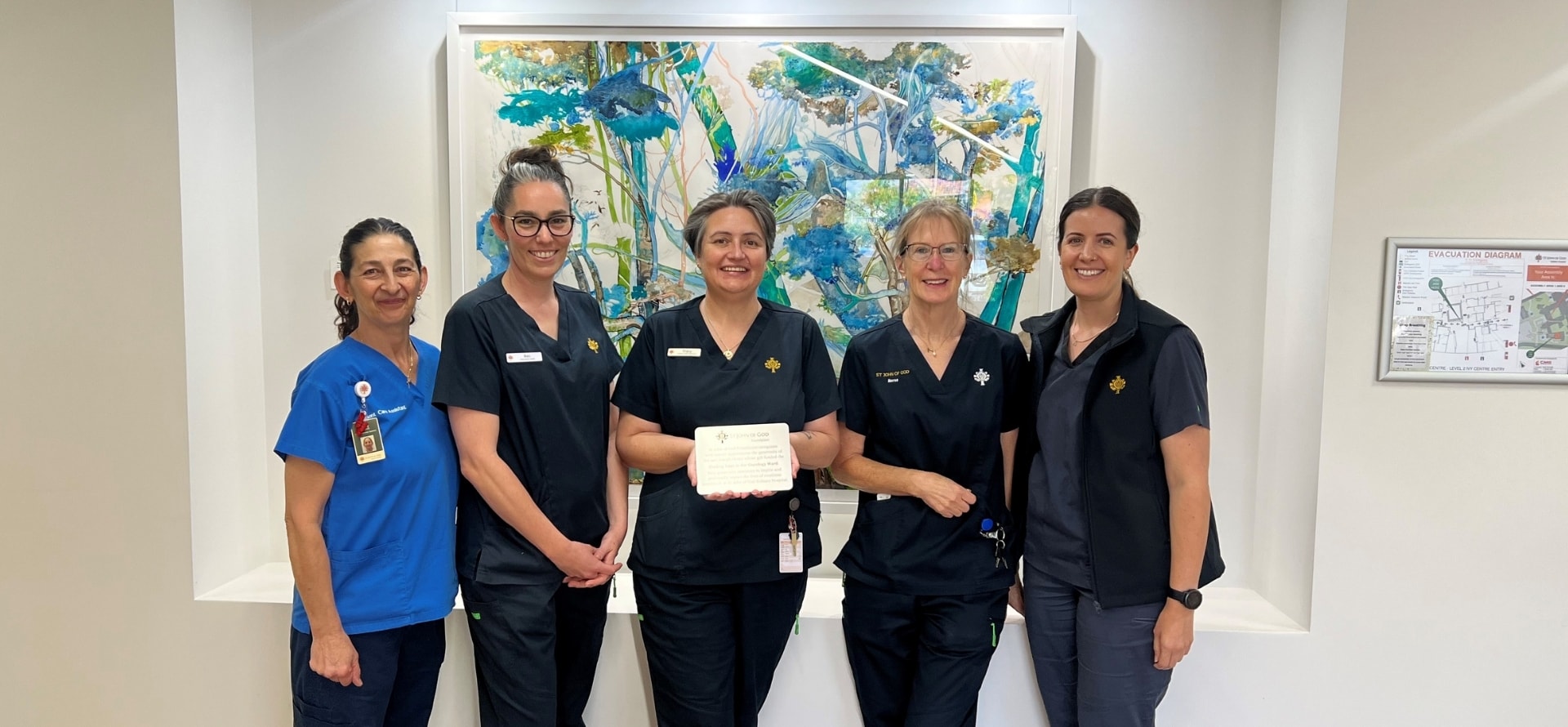 Five women in blue scrubs are smiling while standing before a blue and yellow painting of trees, the middle person showing a small plaque.