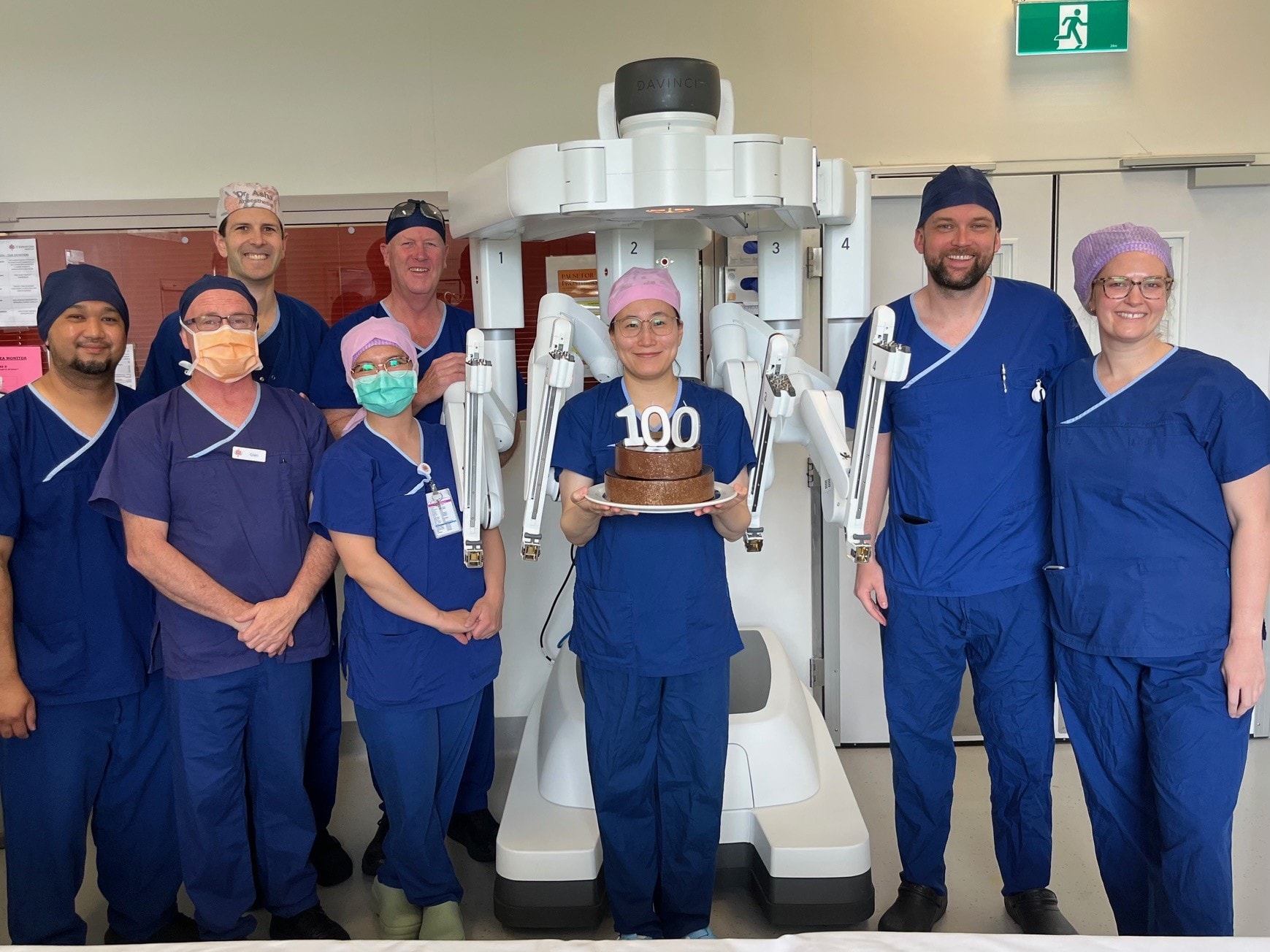 Medical professional in blue scrubs standing in front of a robot with a cake that has the number 100 on top of it
