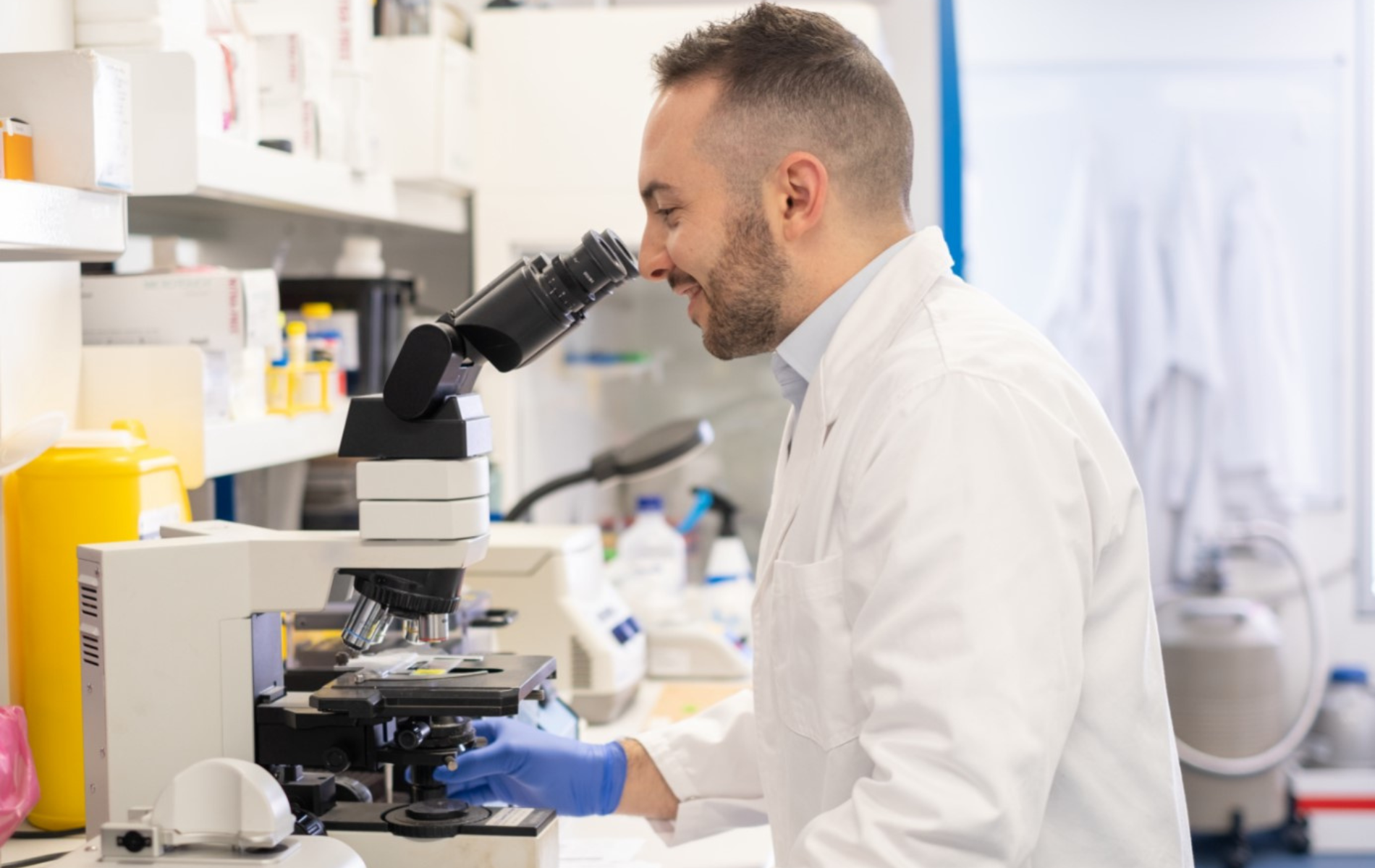 Young man in lab coat, standing in research lab and looking through microscope