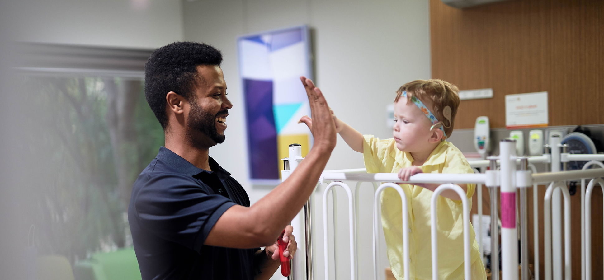 Caregiver smiling and high-fiving with small child, child is in a hospital bed