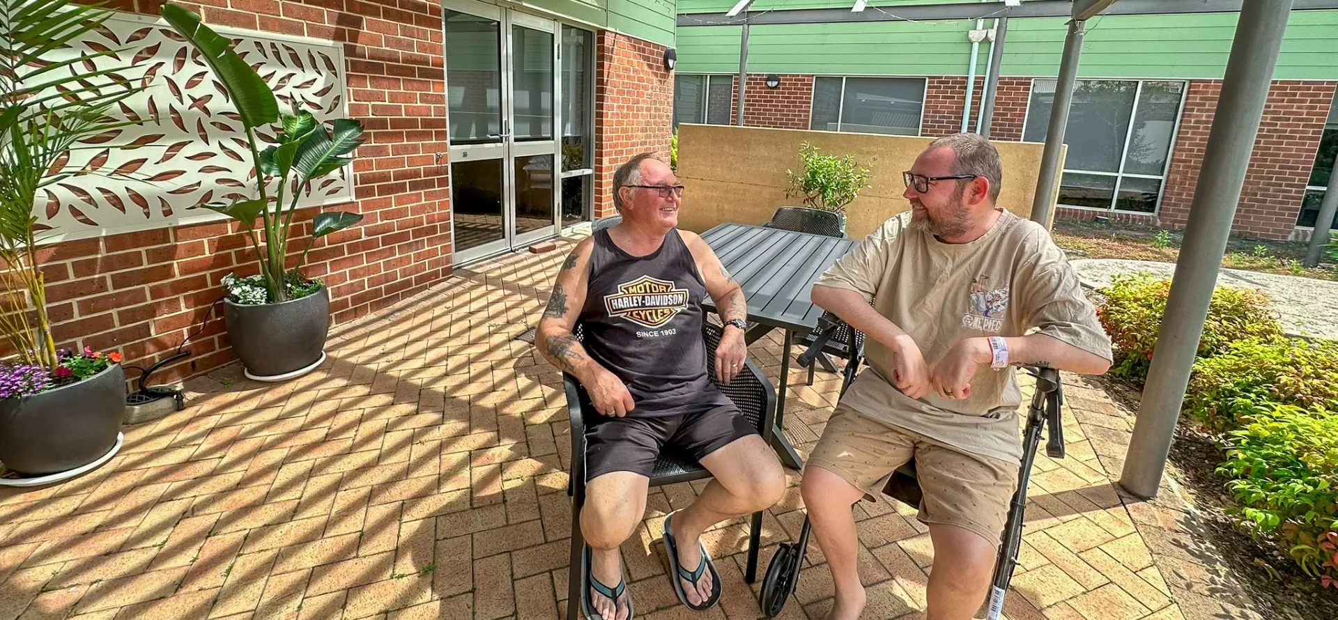 2 elderly men sitting in strollers on the porch of a building while talking to each other.