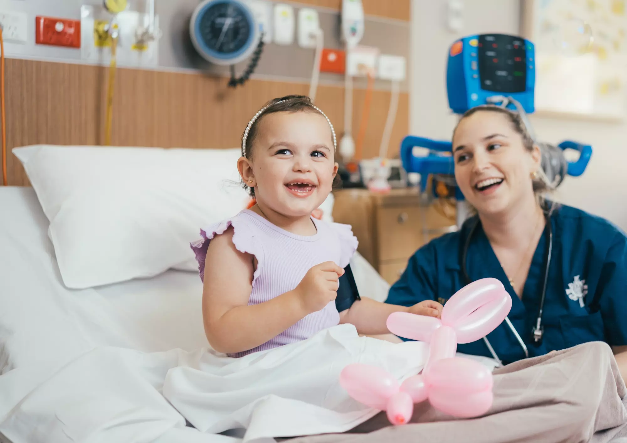 Caregiver and young patient sitting on hospital bed and smiling. The young patient is holding a balloon.