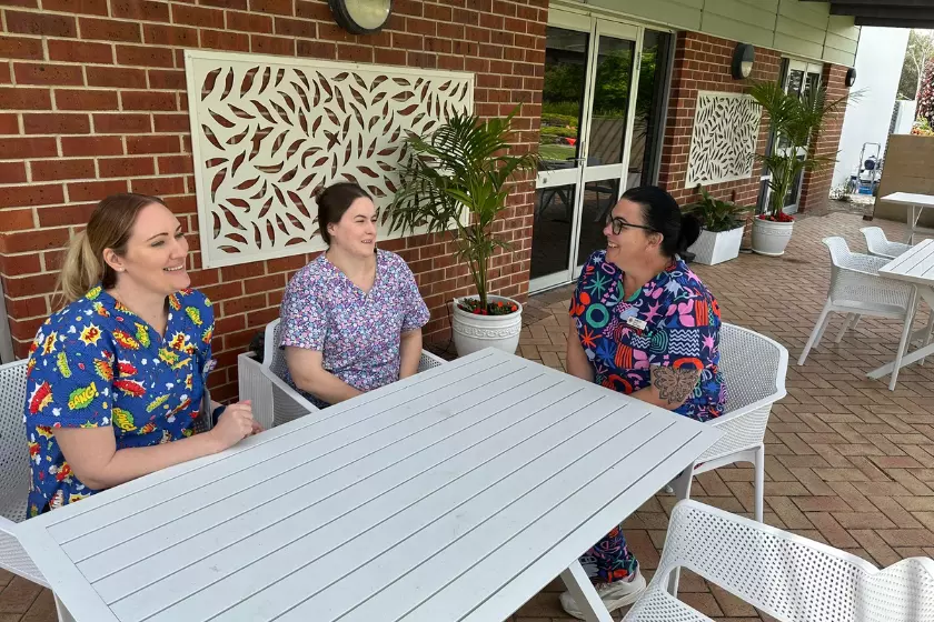 3 ladies in colourful scrubs are talking over a table while sitting outside of a brick building.