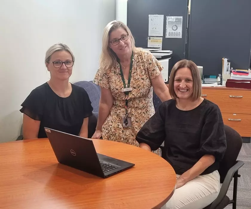 Three people sitting at a desk with an open laptop on it, looking at the camera.