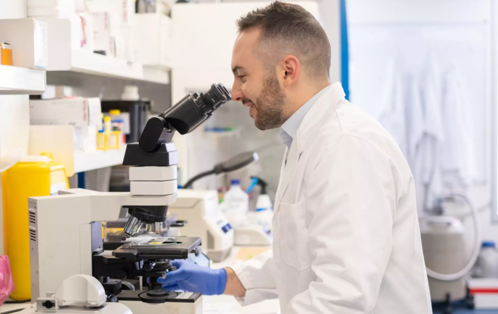 Young man in lab coat, standing in research lab and looking through microscope.