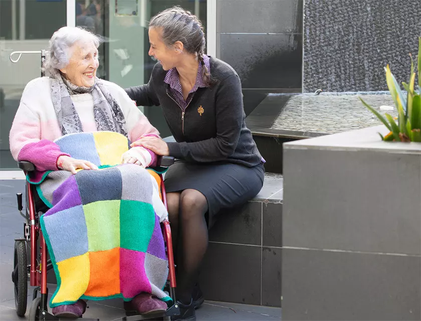 A caregiver sits with an elderly woman in a wheelchair.