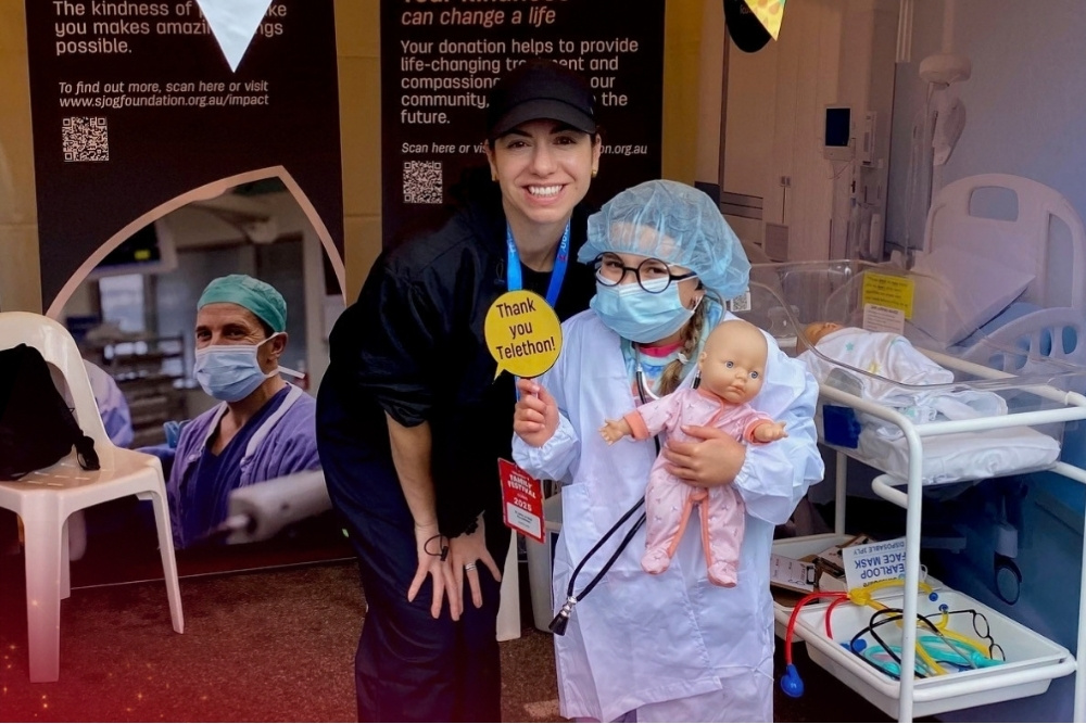 Foundation caregiver smiles with a child playing dress up a medical gown, bonnet and surgical mask